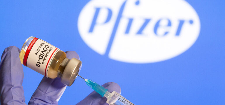 FILE PHOTO: A woman holds a small bottle labeled with a “Coronavirus COVID-19 Vaccine” sticker and a medical syringe in front of displayed Pfizer logo in this illustration