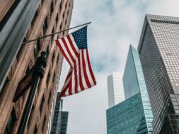 the USA or United States of America flag on a flagpole near skyscrapers under a cloudy sky