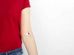 Blood donorship. Young girl in red T-shirt hand taped with patch with red heart after giving blood on gray background. Copy space
