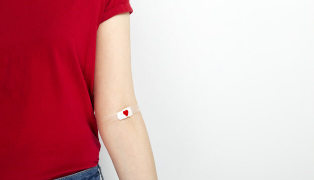 Blood donorship. Young girl in red T-shirt hand taped with patch with red heart after giving blood on gray background. Copy space