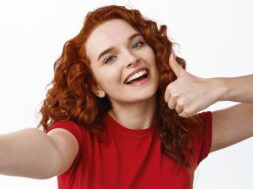Selfie of young happy redhead girl with curly hairstyle showing thumb up, praise and recommend something good, smiling at smartphone camera, white background
