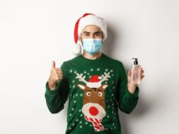 Concept of covid-19 and Christmas holidays. Man recommending hand sanitizer, showing thumb up and antiseptic, wearing medical mask with santa hat, white background