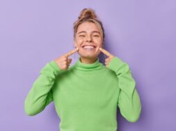 Beautiful happy fair haired European woman points index fingers at mouth forces cheerful smile shows perfect white teeth wears green jumper being in good mood isolated over purple background.