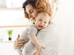 Happy young mom holds precious little child and gently hugging his little body. Kid laughing joyfully and looking in camera with big grey eyes.