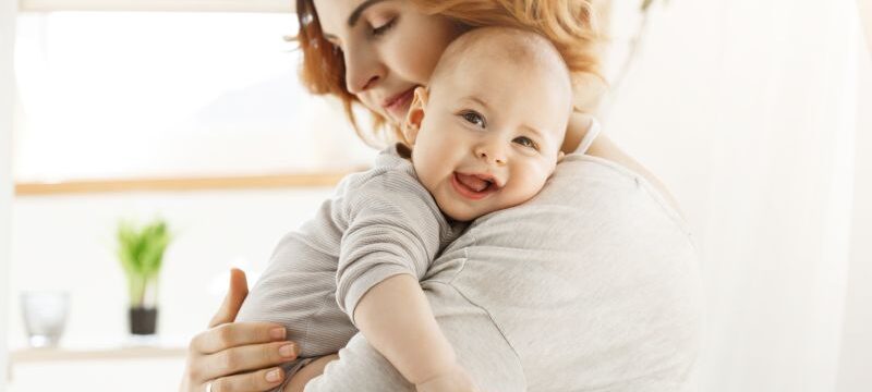 Happy young mom holds precious little child and gently hugging his little body. Kid laughing joyfully and looking in camera with big grey eyes.