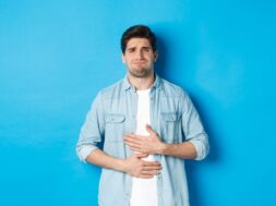 Man holding hands on belly and grimacing from pain, complaining on stomach ache, standing against blue background