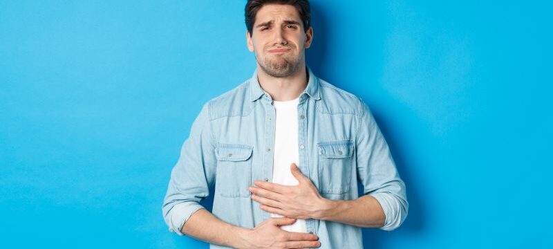 Man holding hands on belly and grimacing from pain, complaining on stomach ache, standing against blue background