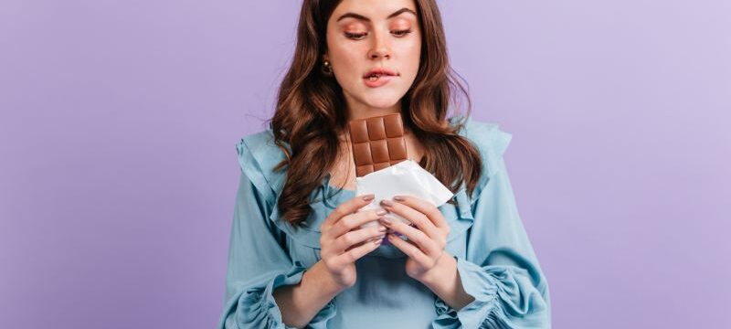 Portrait of funny brunette woman biting her lip in anticipation of tasty lunch. Girl in blue dress looks at delicious chocolate.