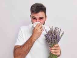 Indoor shot of allergic man blows nose in handkerchief has sneezing and rhinitis allergy to lavender red swelling eyes suffers from unpleasant symptoms isolated on white wall. Sickness concept