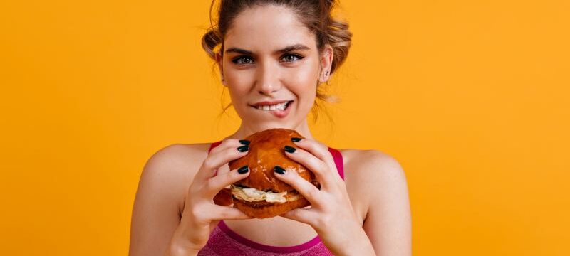 Adorable young lady eating burger. Indoor portrait of girl with big sandwich