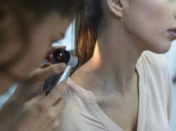Dermatologist in latex gloves holding dermatoscope while examining attractive patient with skin disease. Female dermatologist examining patient with dermascope, looking for signs of skin cancer.
