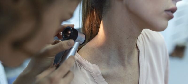 Dermatologist in latex gloves holding dermatoscope while examining attractive patient with skin disease. Female dermatologist examining patient with dermascope, looking for signs of skin cancer.