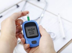 Hand holding a blood glucose meter measuring blood sugar, the background is a stethoscope and chart file