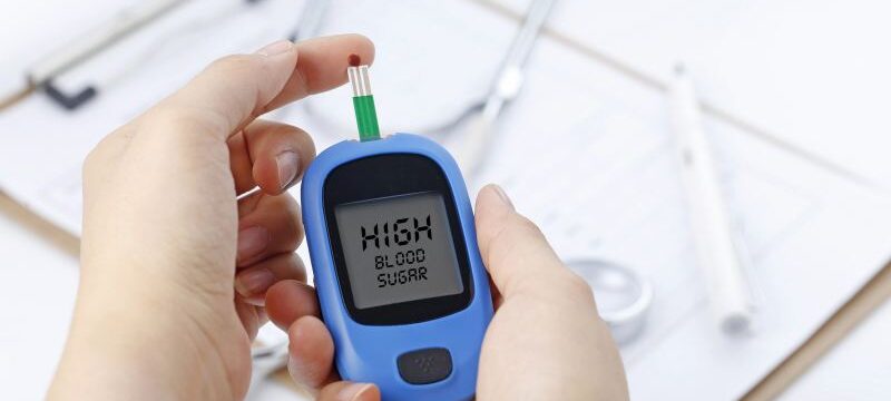 Hand holding a blood glucose meter measuring blood sugar, the background is a stethoscope and chart file