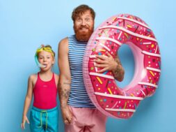 Indoor shot of delighted caring father in striped vest and shorts, holds inflated swimring, touches hand of little daughter in snorkel mask, have fun in water park, enjoy bathing, stand over blue wall