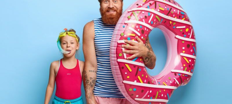 Indoor shot of delighted caring father in striped vest and shorts, holds inflated swimring, touches hand of little daughter in snorkel mask, have fun in water park, enjoy bathing, stand over blue wall