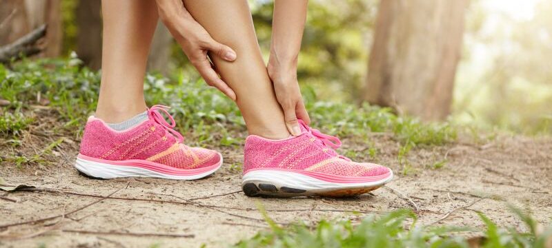 Running sport injury. Female athlete jogger wearing pink sneakers touching her twisted or sprained ankle while jogging or running outdoors. Woman runner massaging her calf muscle before workout