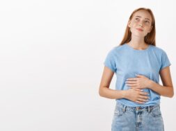 Woman remembering tasty food she ate in new restaurant rubbing tummy or belly looking at upper left corner nostalgic and happy liking eat delicious meals standing in blue t-shirt over gray background