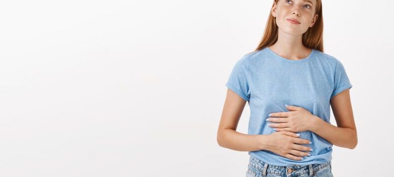 Woman remembering tasty food she ate in new restaurant rubbing tummy or belly looking at upper left corner nostalgic and happy liking eat delicious meals standing in blue t-shirt over gray background