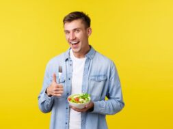 Healthy lifestyle, people and food concept. Happy smiling man showing thumbs-up satisfied with delicious breakfast, eating salad, being on diet, trying stay fit, standing yellow background