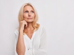 Thoughtful blonde middle aged woman ponders on something keeps hand near face has healthy skin minimal makeup makes choice wears white blouse poses indoor blank copy space for your promotion