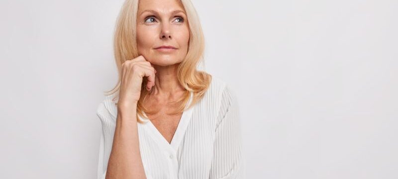 Thoughtful blonde middle aged woman ponders on something keeps hand near face has healthy skin minimal makeup makes choice wears white blouse poses indoor blank copy space for your promotion
