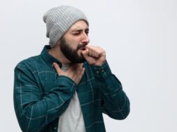 unhealthy young man wearing winter hat keeping hand on chest and fist in front of mouth coughing with closed eyes isolated on white background with copy space