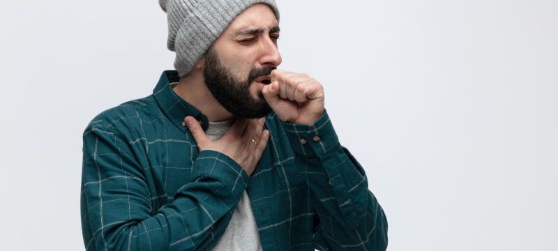 unhealthy young man wearing winter hat keeping hand on chest and fist in front of mouth coughing with closed eyes isolated on white background with copy space
