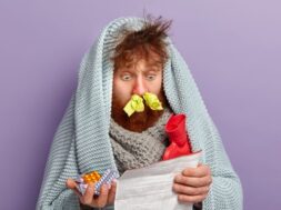 Healthcare concept. Unhealthy stupefied foxy man stares at medical leaflet, surprised with side effect of tablets, warms with coverlet and hot water bag, cures fever and cold, has tissues in nostrils
