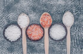 Top view sea salt in bowls and wooden spoons with himalayan salt on gray textured background. vertical