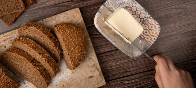 top-view-woman-hand-cutting-butter-with-knife-sliced-sandwich-bread-cutting-board-wooden-background_141793-6005