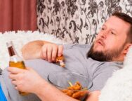 fat man drinks beer and eats unhealthy food chicken wings, bored in front of the TV outlook on the couch. The concept of malnutrition, quarantine at home, alcoholism