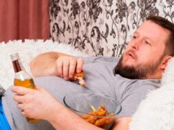 fat man drinks beer and eats unhealthy food chicken wings, bored in front of the TV outlook on the couch. The concept of malnutrition, quarantine at home, alcoholism