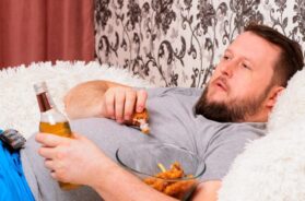 fat man drinks beer and eats unhealthy food chicken wings, bored in front of the TV outlook on the couch. The concept of malnutrition, quarantine at home, alcoholism