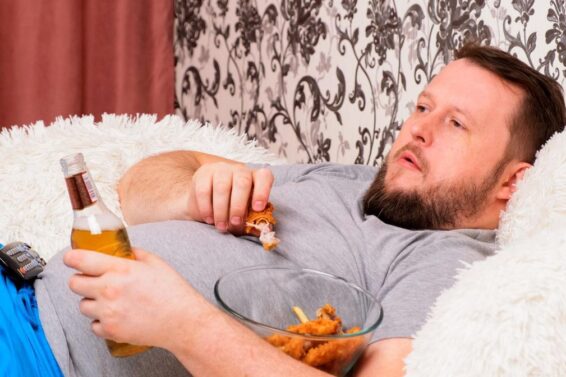 fat man drinks beer and eats unhealthy food chicken wings, bored in front of the TV outlook on the couch. The concept of malnutrition, quarantine at home, alcoholism