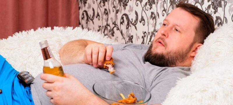 fat man drinks beer and eats unhealthy food chicken wings, bored in front of the TV outlook on the couch. The concept of malnutrition, quarantine at home, alcoholism