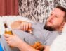 fat man drinks beer and eats unhealthy food chicken wings, bored in front of the TV outlook on the couch. The concept of malnutrition, quarantine at home, alcoholism